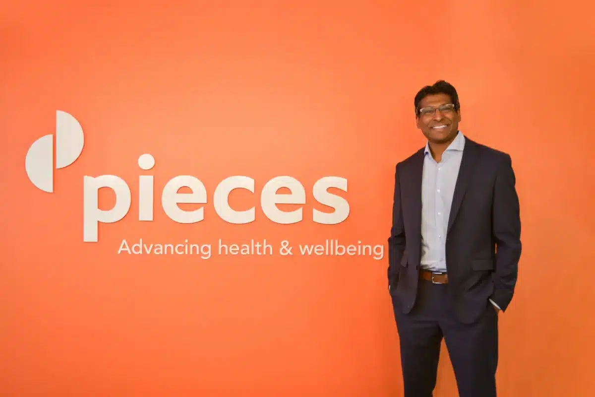A professionally dressed man with dark skin, short black hair with gray streaks, and glasses stands smiling in front of a bright orange wall. He wears a dark blue business suit and light blue shirt. To his left, large white text reads “pieces – Advancing health & wellbeing.”