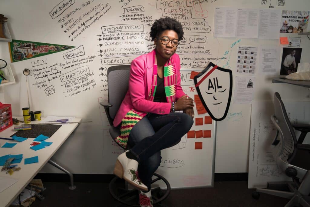 Dr. Joy Buolamwini seated on a chair in front of a whiteboard filled with handwritten notes and diagrams on systemic bias, healthcare, and inclusion. She is wearing a pink blazer, a multicolored kente scarf, and white boots with floral designs.