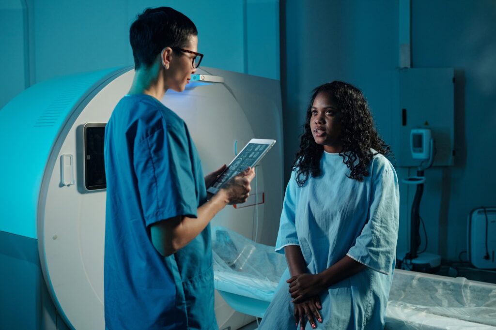 A female patient in a medical gown sits on the edge of an MRI scanner table while a healthcare professional holds a digital tablet showing scan images. This clinical setting represents diagnostic workflows supported by MedGemma models in modern healthcare.