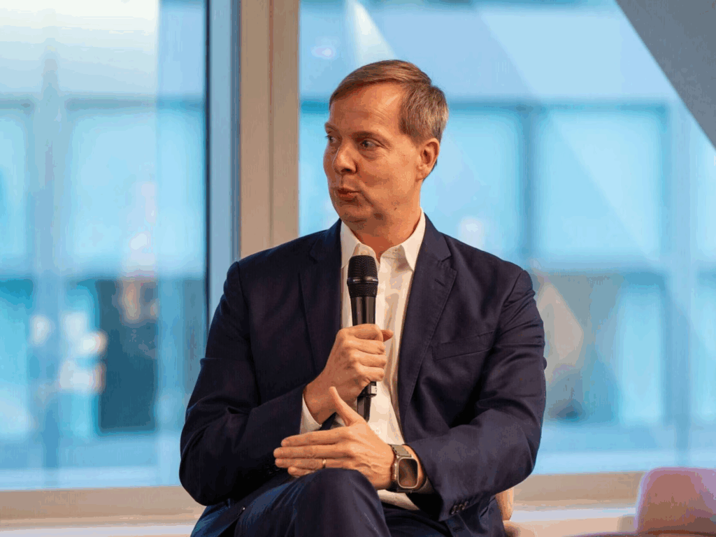 A man in a navy suit holding a microphone, seated and speaking during a panel discussion, with a blurred glass window and modern office background.