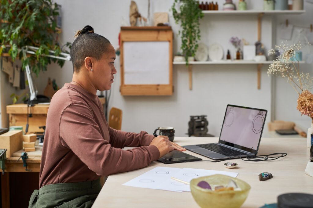 Person using a digital drawing tablet and laptop to create AI jewelry design prototypes in a modern studio workspace.